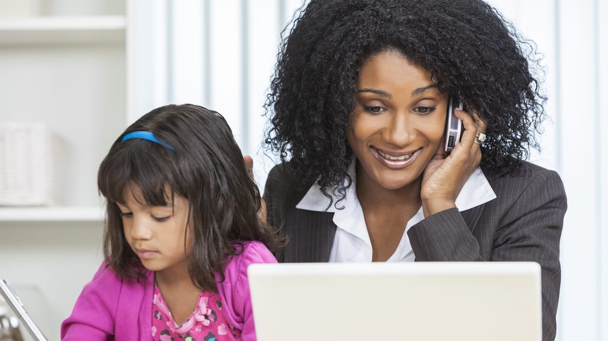 An woman on a cell phone with a young girl in front of a laptop.