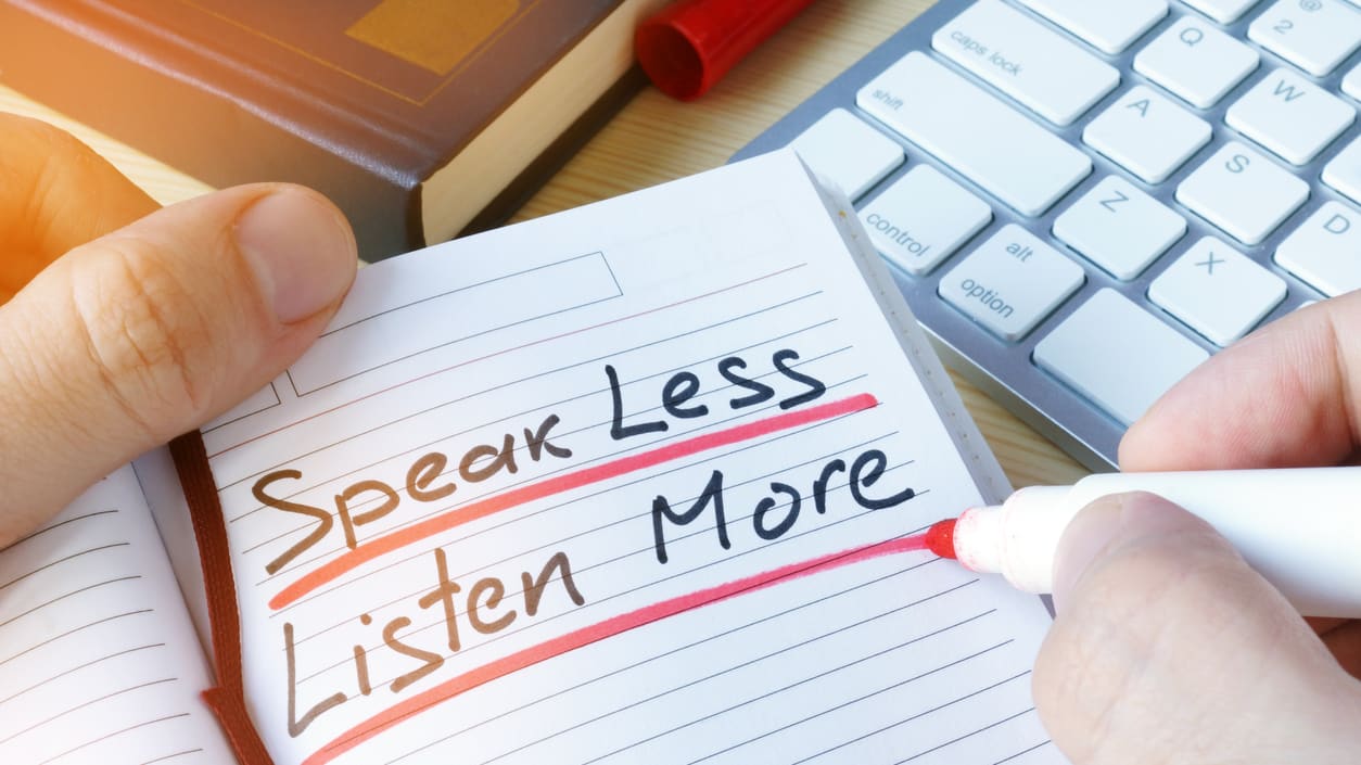 A person writing the words speak less listen more on a notebook.
