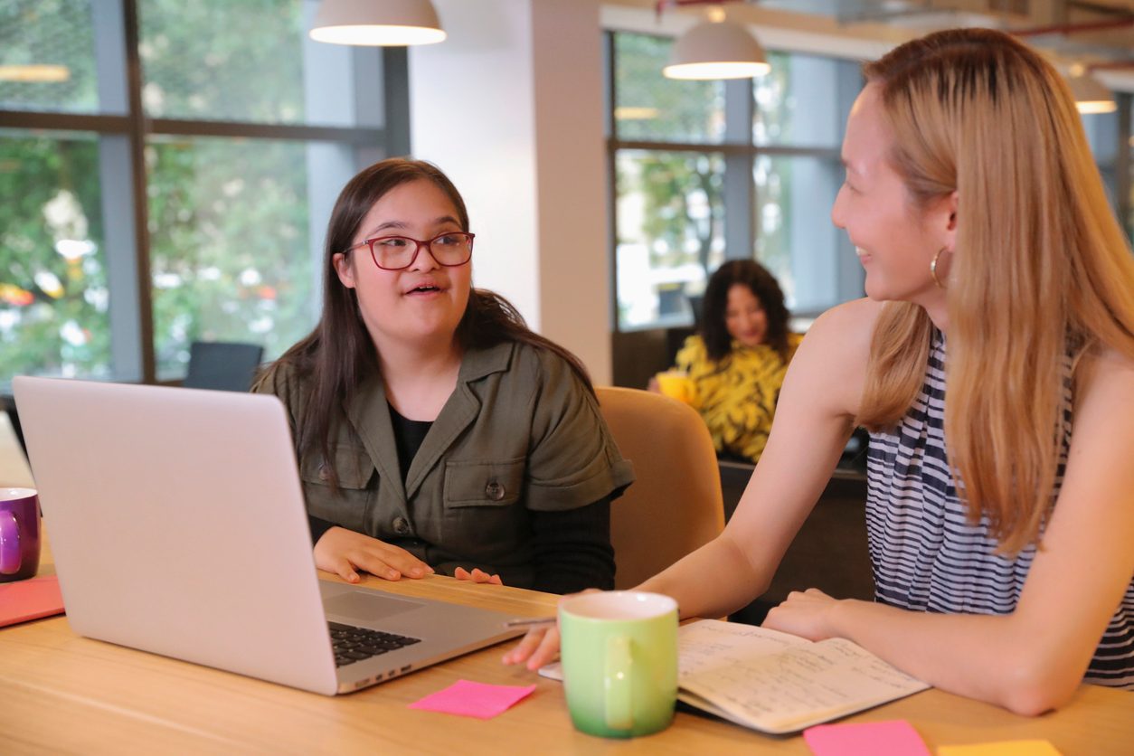 Two women sit at a light wooden table in a bright, modern indoor space, engaged in a friendly conversation. The woman on the left, with long dark brown hair and red-framed glasses, appears to have Down syndrome and is speaking or reacting while resting her hands near an open laptop. The woman on the right, with long blonde hair and a striped sleeveless top, smiles warmly, her hand resting near a green mug and an open notebook with handwritten notes. The table also features sticky notes, a purple mug, and other work items. In the blurred background, a third woman with curly hair and a yellow patterned top sits at another table, smiling. Large windows let in natural light, creating an open, inclusive, and collaborative atmosphere.