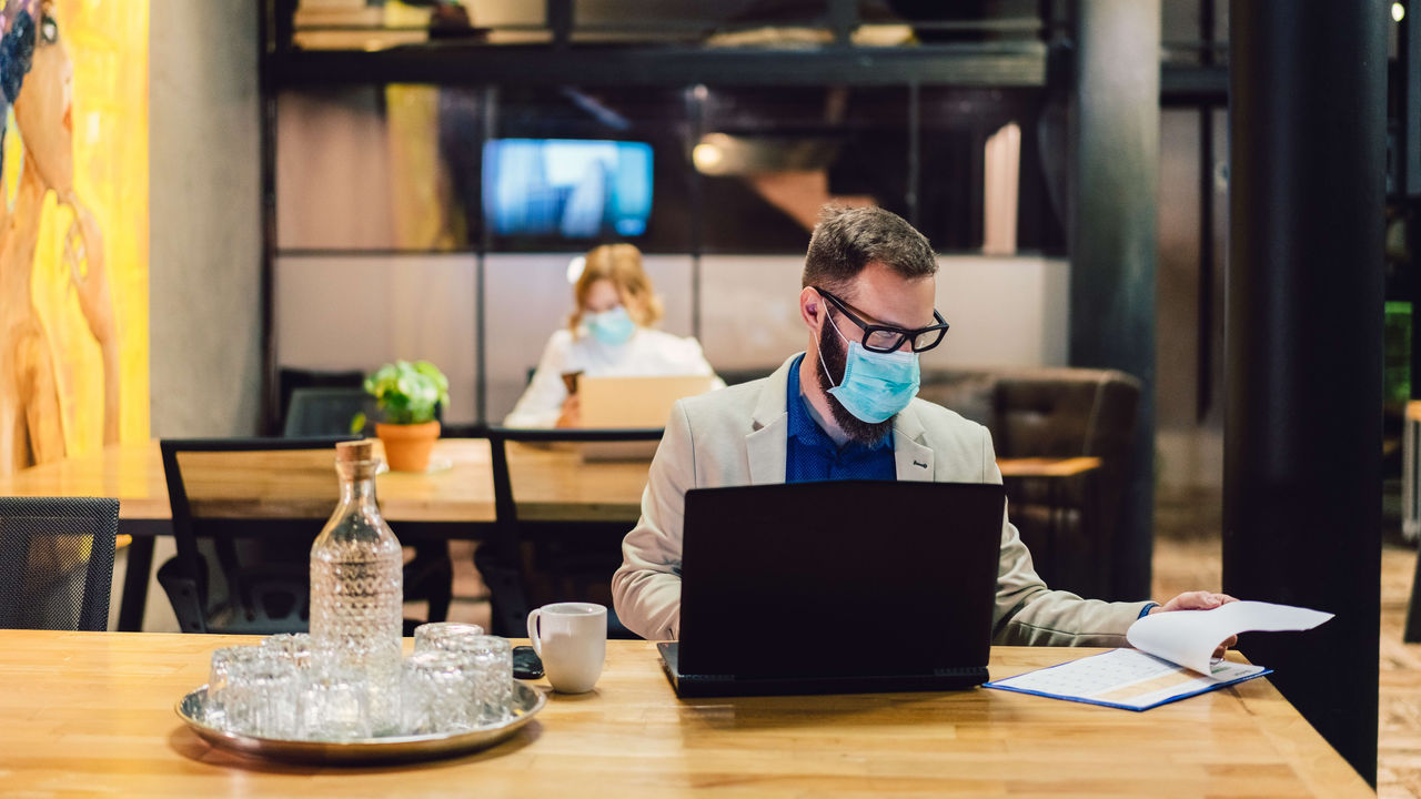 A man wearing a face mask sits at a table with a laptop.