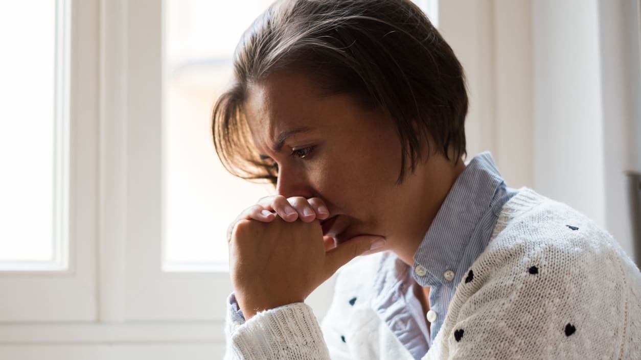 A woman is sitting in front of a window with her hands on her chin.