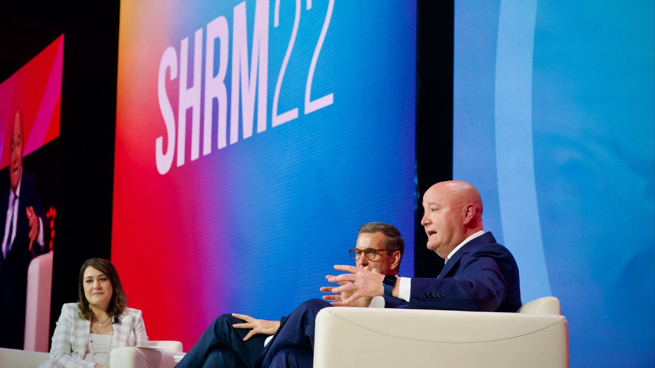 Three people sitting on chairs at a conference with a shrm2 sign in the background.
