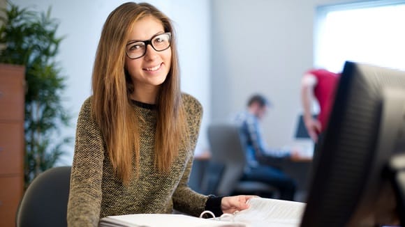 A young woman in glasses sitting at a desk in an office.