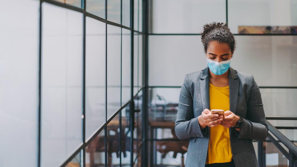 A woman wearing a surgical mask is using her phone in an office.