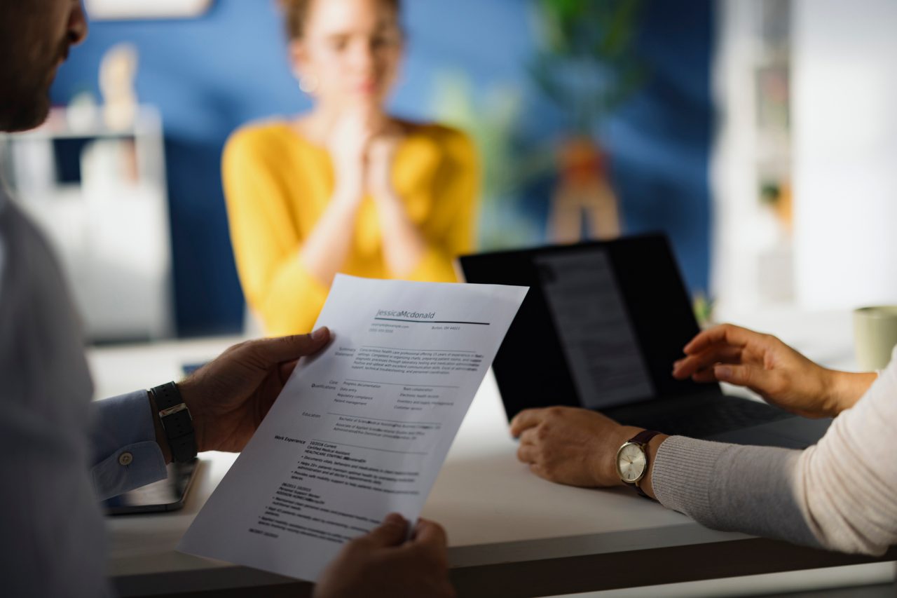 Three people sit at a table during a job interview; one person holds a resume, another uses a laptop, and a third person sits across from them with hands clasped.
