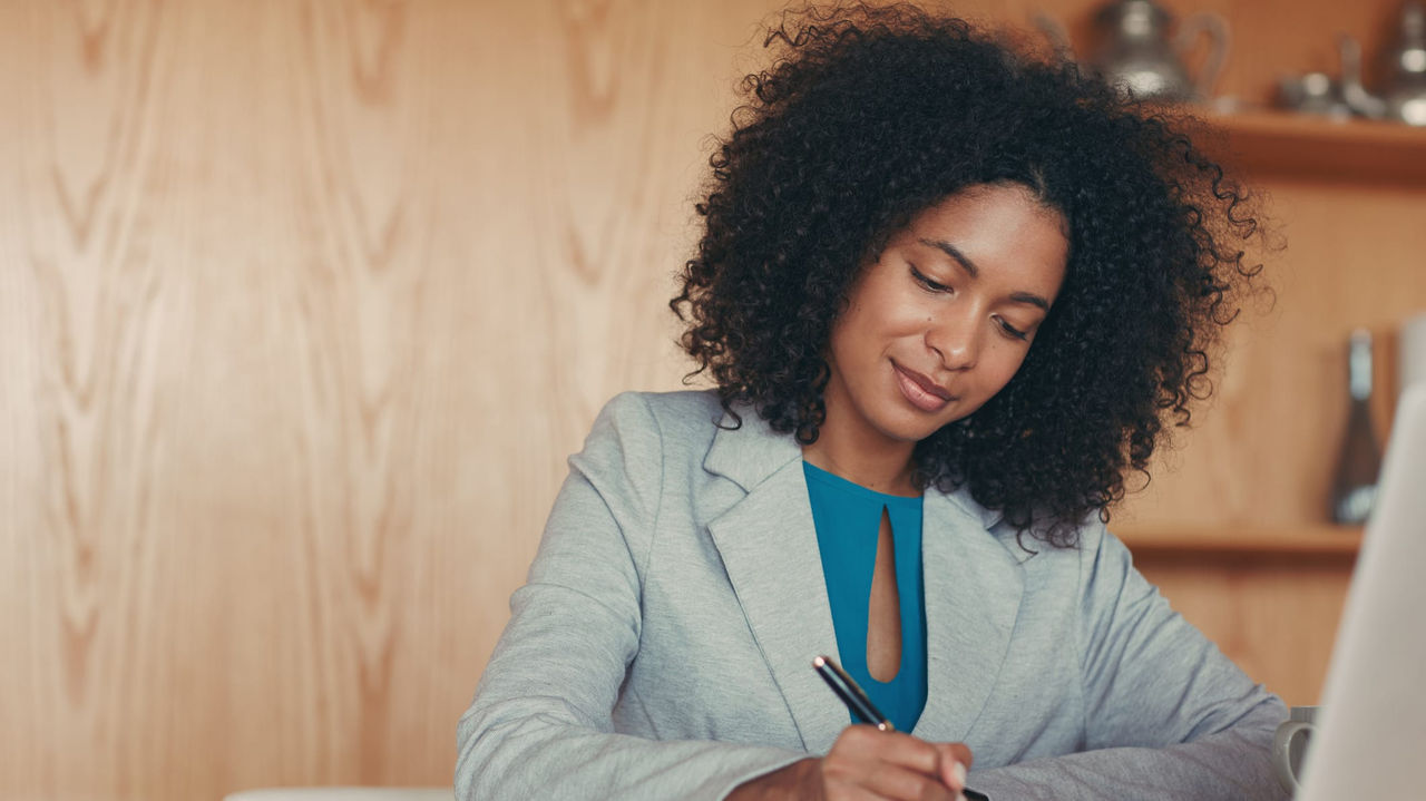 A woman writing in a notebook while sitting at a desk with a laptop.