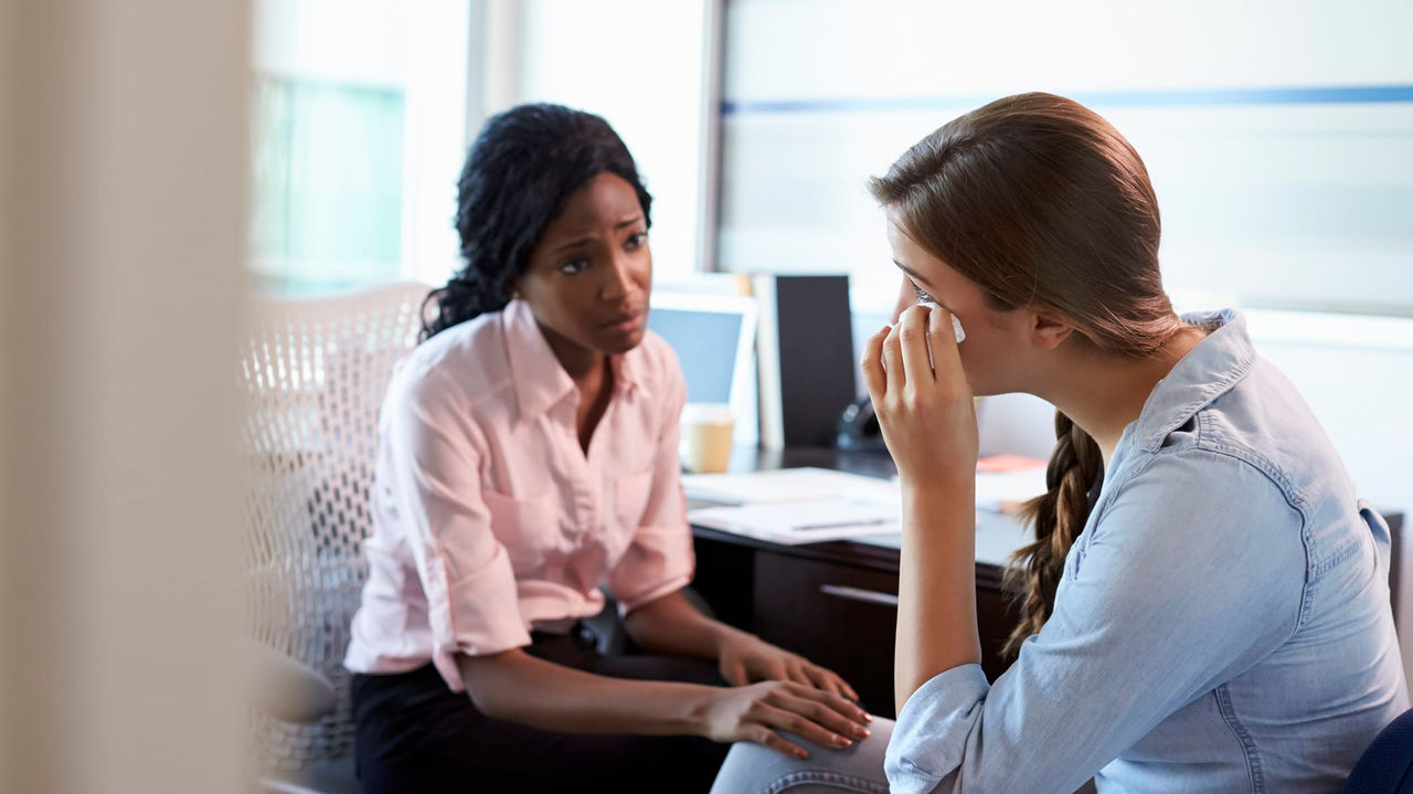 Two women sitting in an office talking to each other.