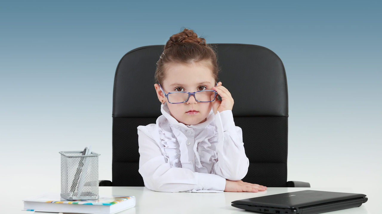 A little girl sitting at a desk with glasses on.