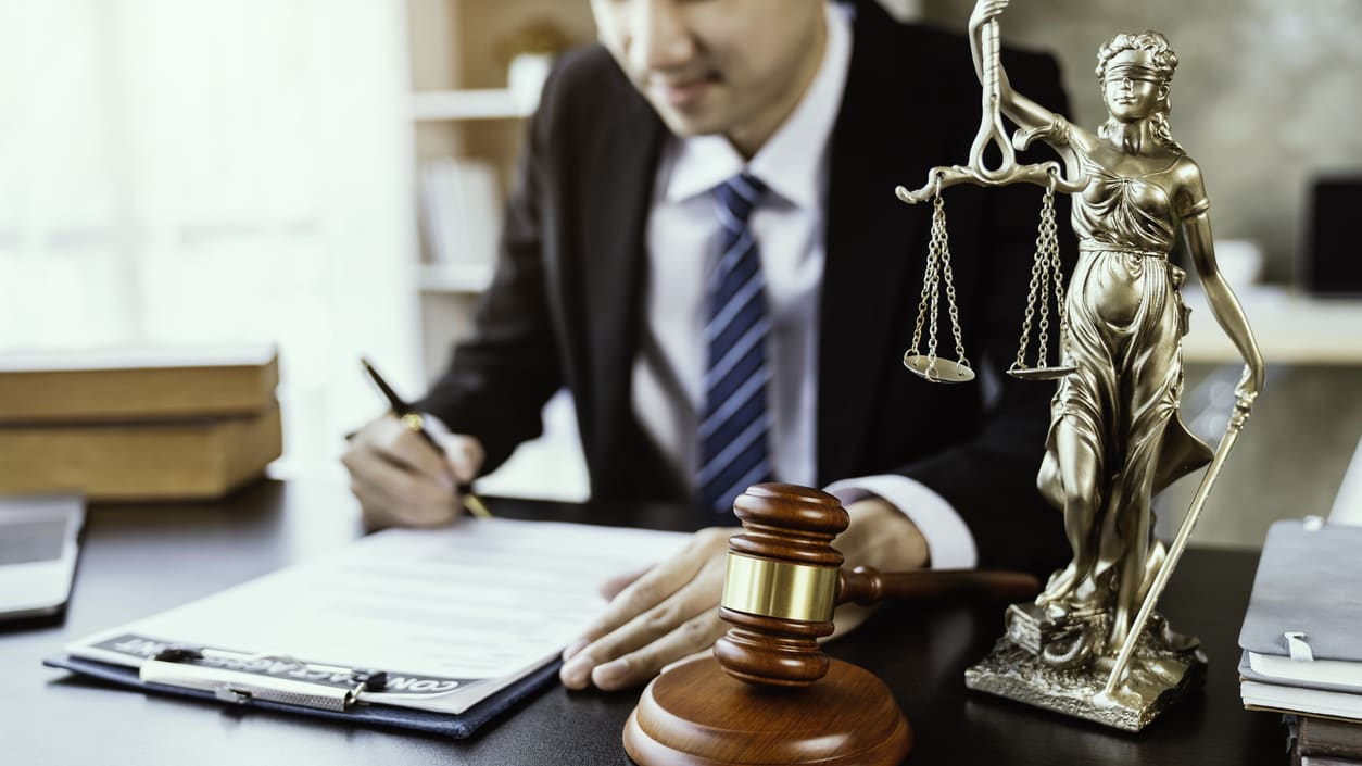 A man in a suit sitting at a desk with a statue of justice.