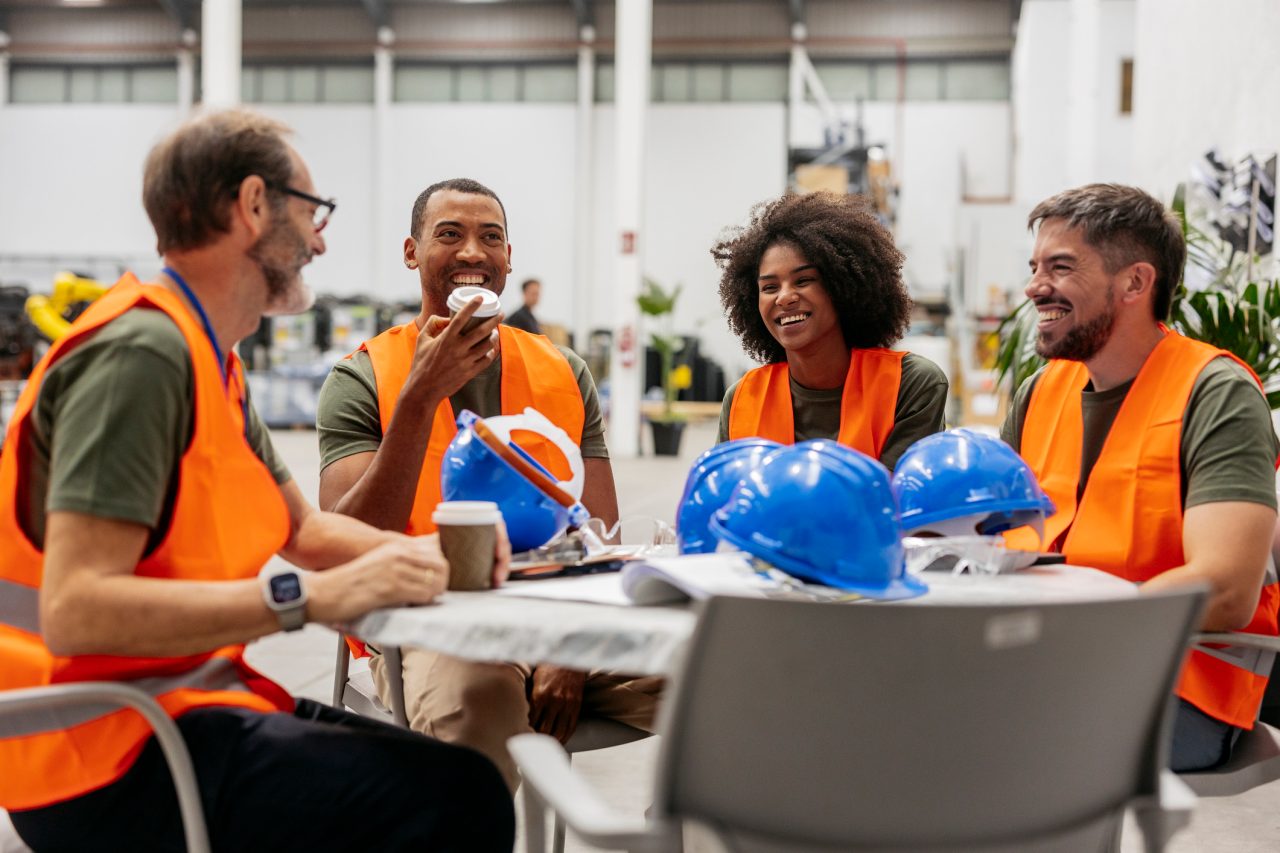 Four diverse employees wearing orange safety vests sit around a table. 