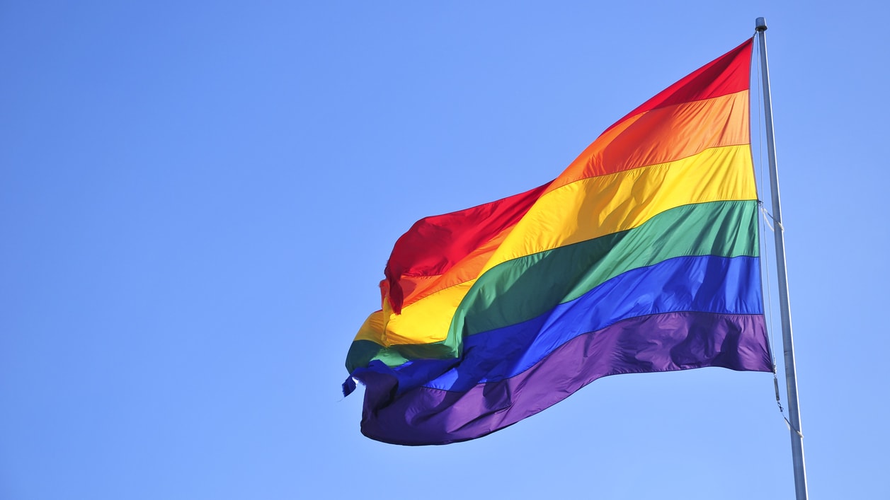 A rainbow flag flying in the wind against a blue sky.