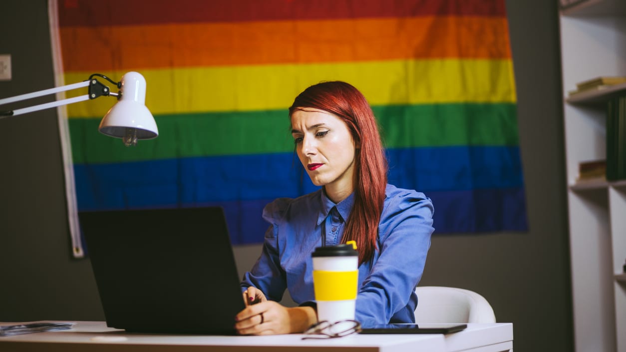 A woman working on a laptop in front of a rainbow flag.