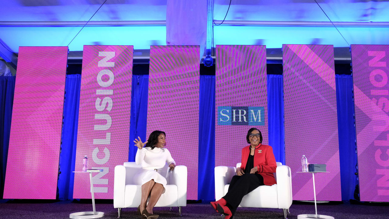 Two women sitting in chairs on a stage with a pink background.