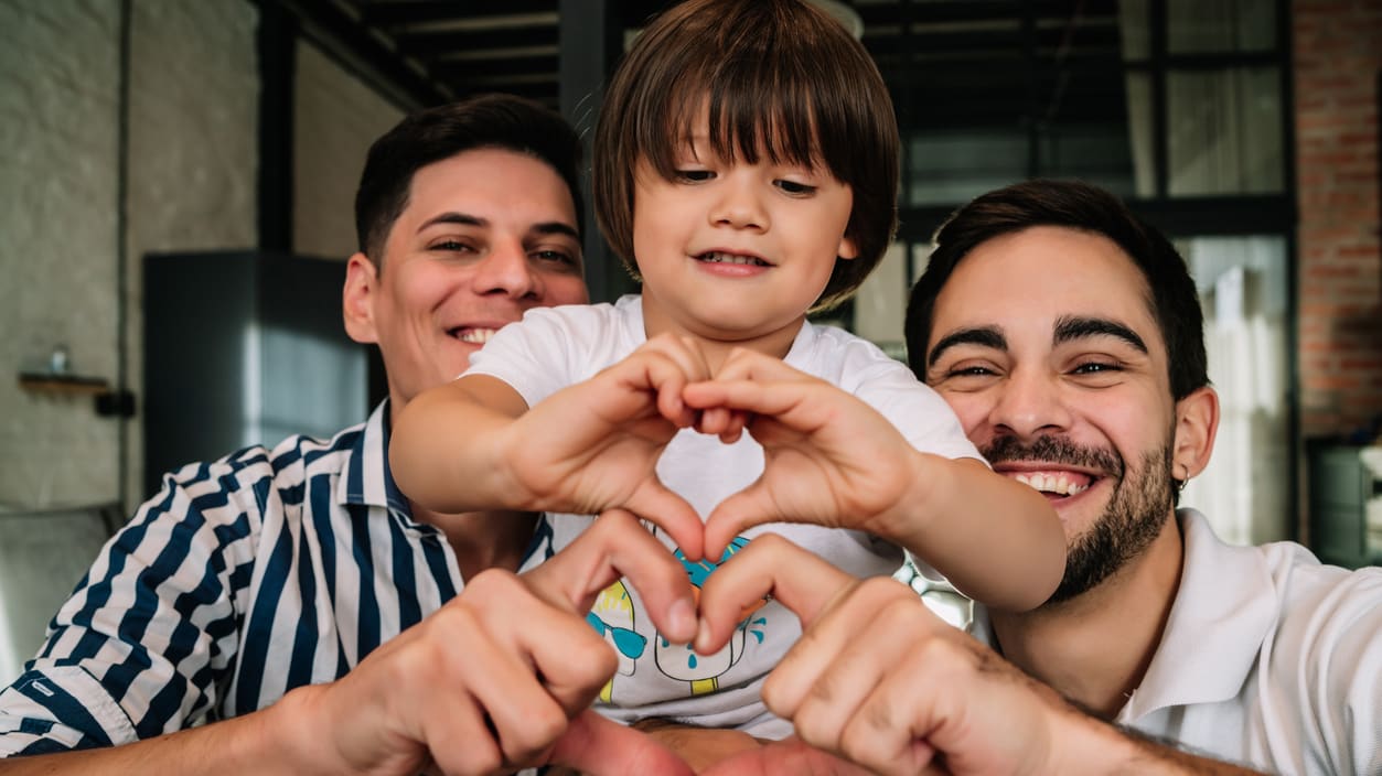 Two dads and their son making a heart shape with their hands.