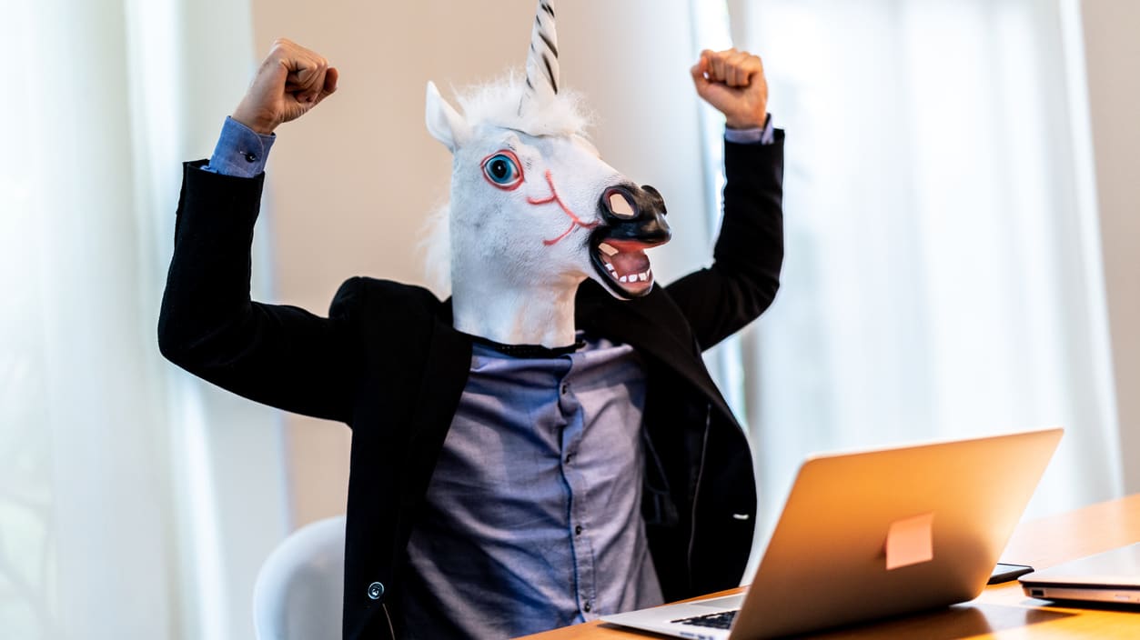 A businessman wearing a unicorn mask in front of a laptop.
