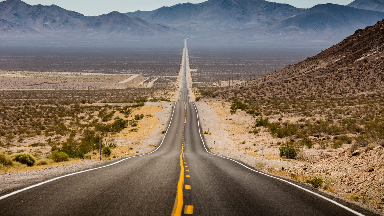 A long road in the desert with mountains in the background.