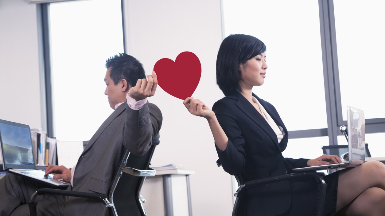 A man and woman in an office holding a red paper heart.