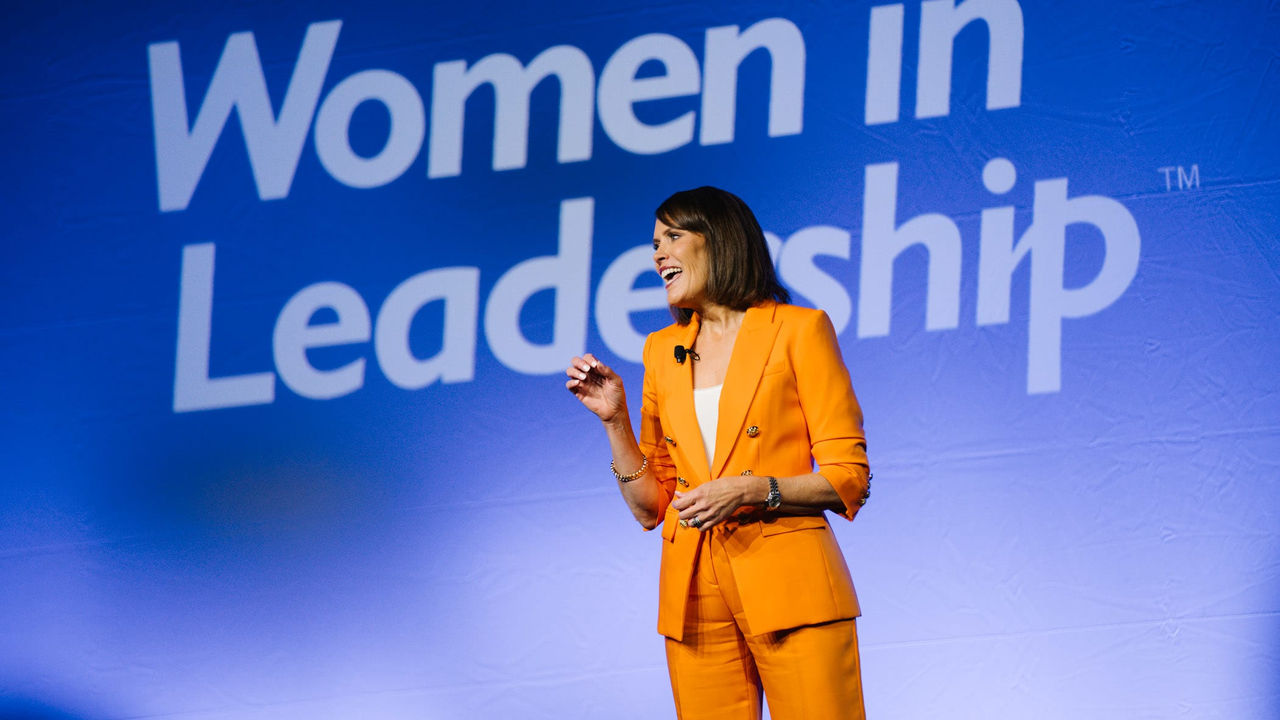 A woman in orange standing in front of a sign that says women in leadership.