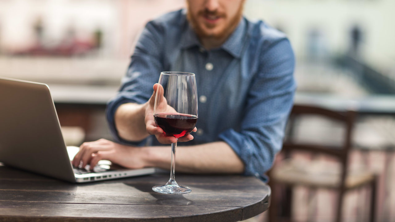 A man holding a glass of wine while working on his laptop.