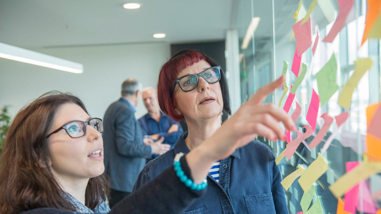 Two women looking at sticky notes on a wall.