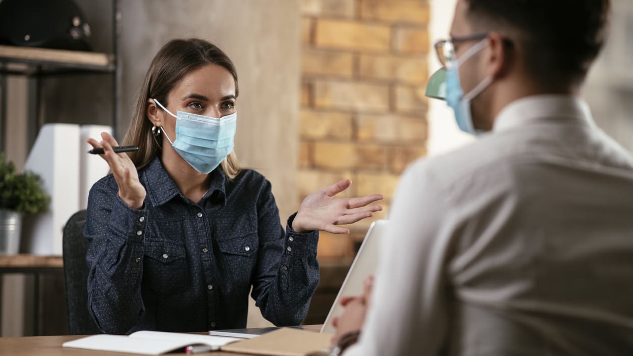 A woman wearing a surgical mask is talking to a man at a desk.
