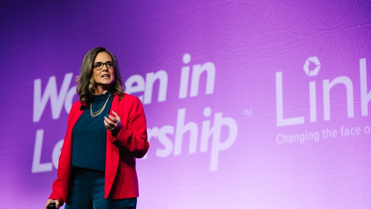 A woman standing in front of a purple screen with the words women in leadership.
