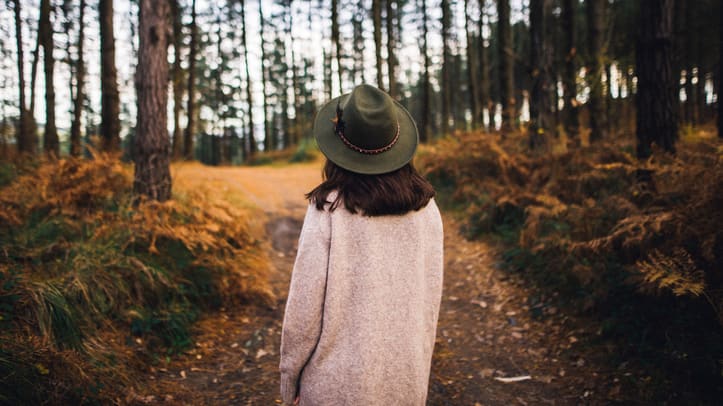 A woman wearing a hat walks down a path in the woods.