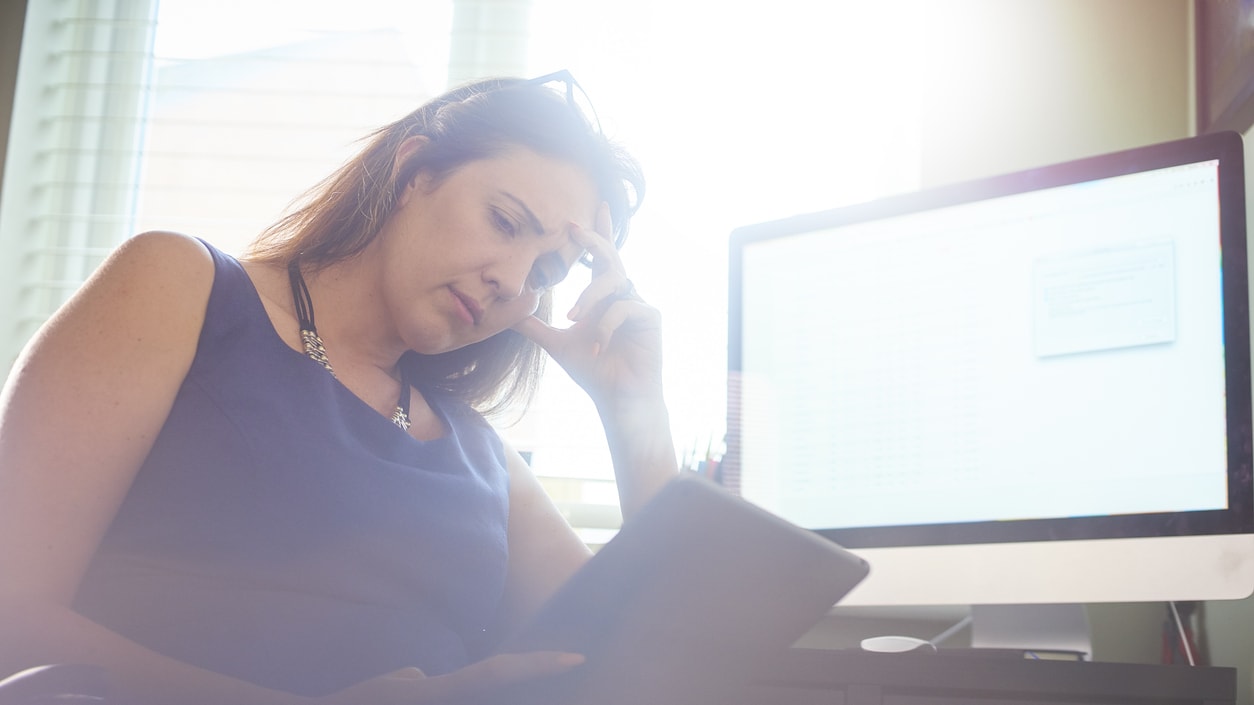 A woman sitting in front of a computer looking at a tablet.