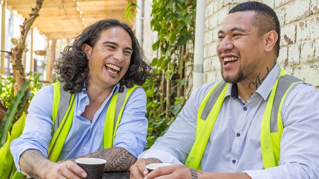 Two men in safety vests laughing at an outdoor table.