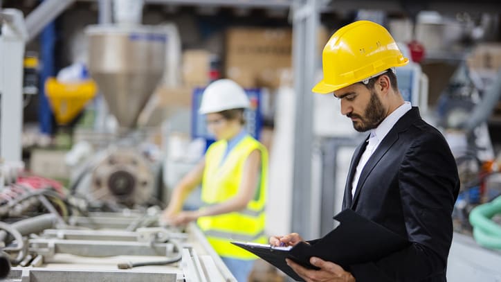 A man in a hard hat is holding a clipboard in a factory.