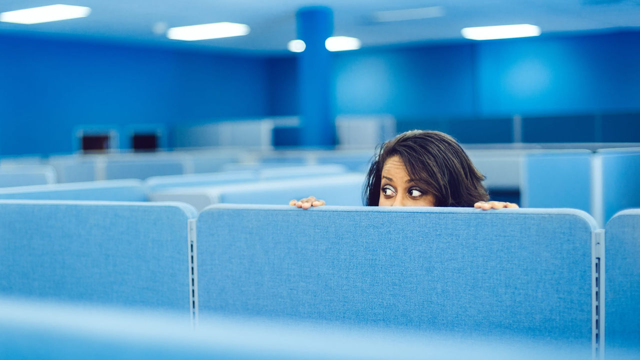 A woman peeking out from behind a blue cubicle.
