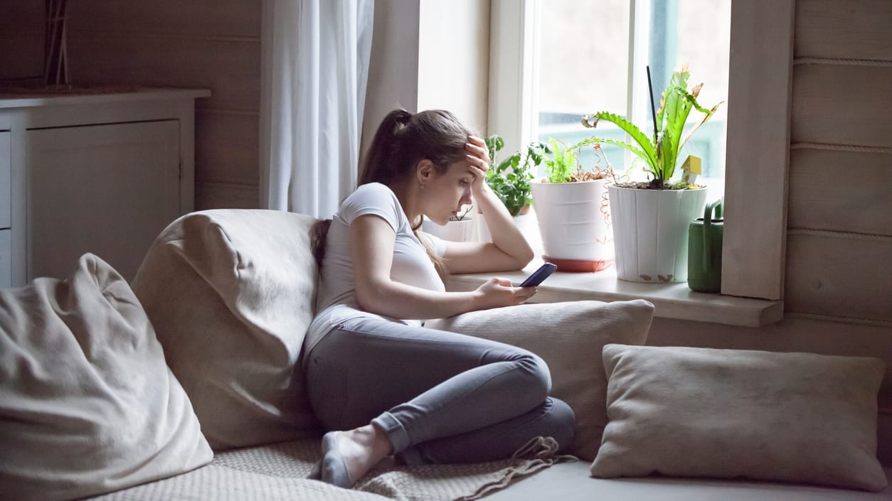 A woman sitting on a couch looking at her phone by a window.