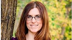 A woman wearing glasses and a purple shirt is leaning against a tree.