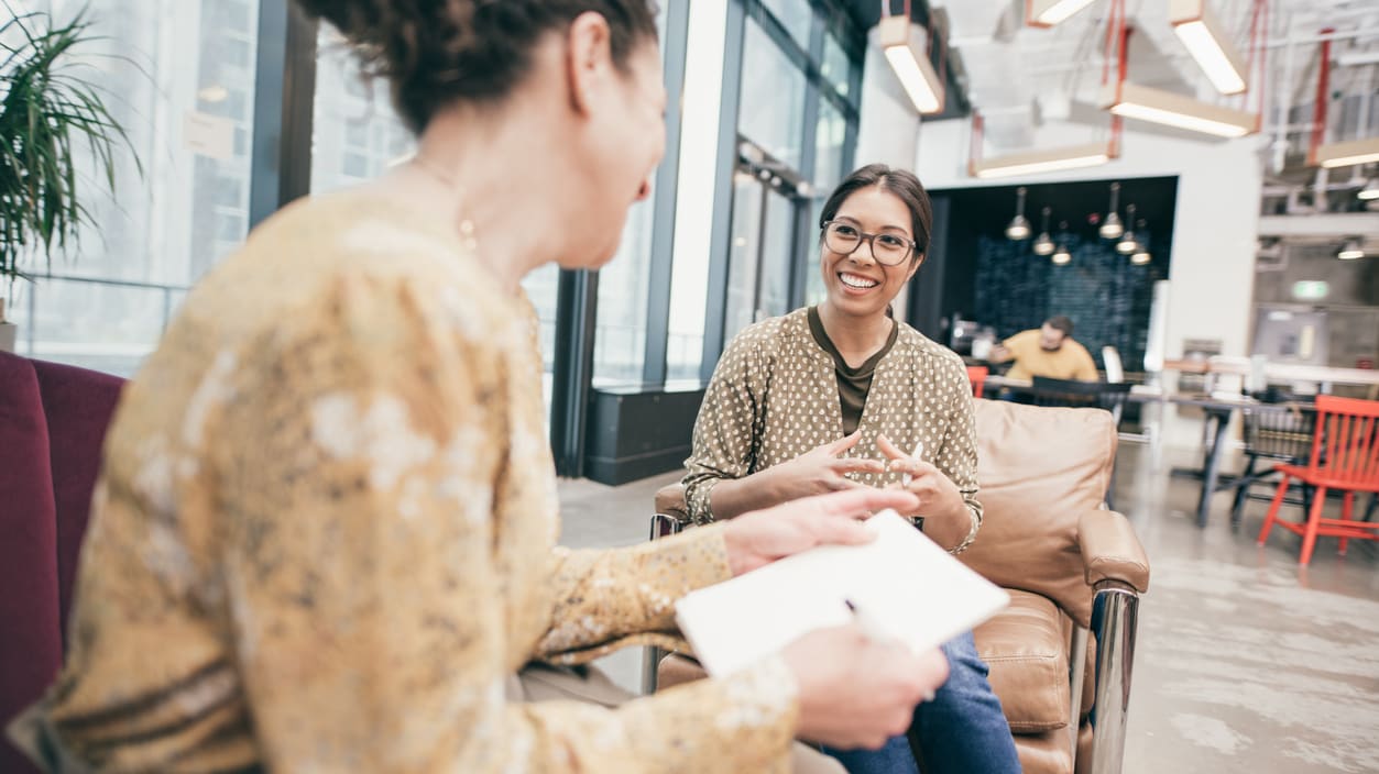 Two women sitting in an office talking to each other.