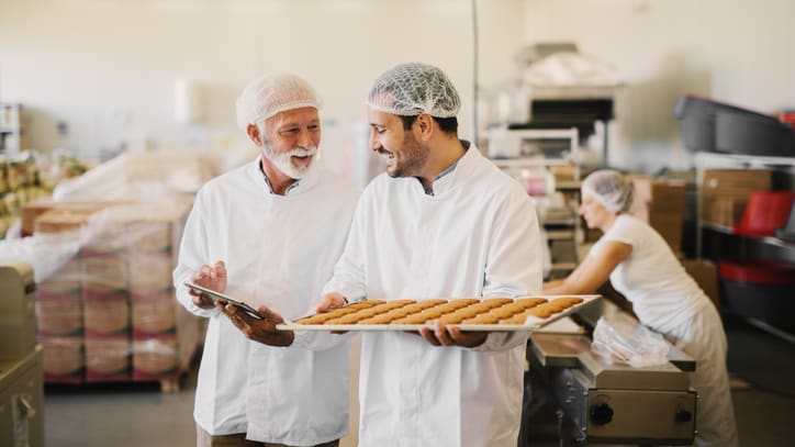 Two men in a factory holding a tray of doughnuts.