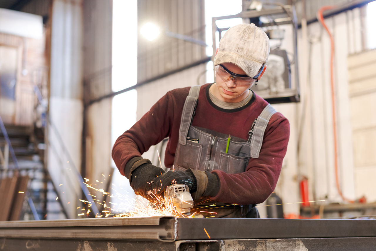 Focused young tradesman in protective eyewear using angle grinder creating sparks in manufacturing facility