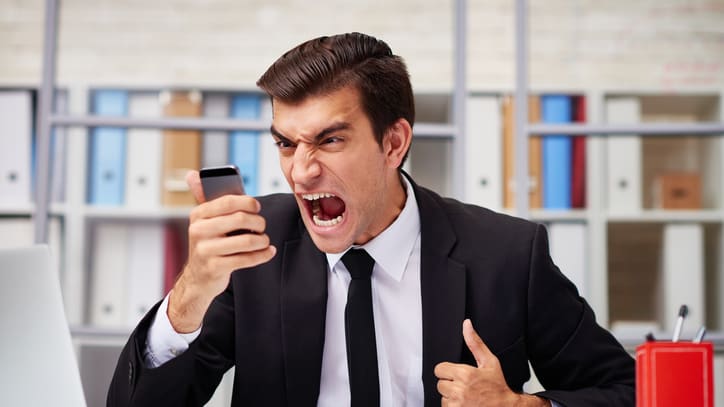 A businessman yelling at his phone while sitting at his desk.