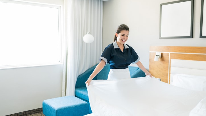 A maid in a hotel room putting a sheet on a bed.