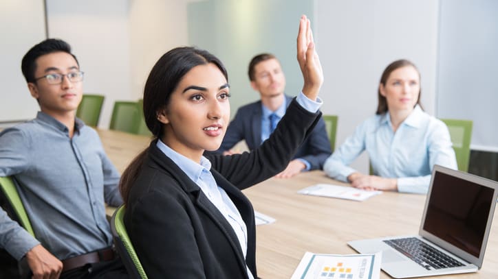 A group of business people in a meeting room raising their hands.