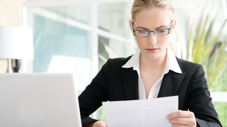 A business woman is looking at a piece of paper in front of a laptop.
