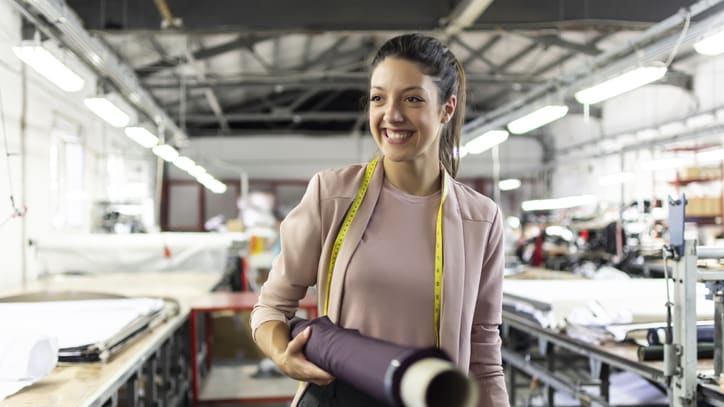 A woman is standing in a factory holding a roll of fabric.