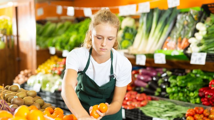 A woman in a green apron picking out fruit in a grocery store.