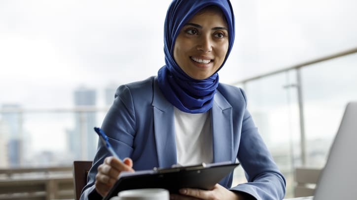 A muslim woman in a hijab sitting at a table with a laptop.