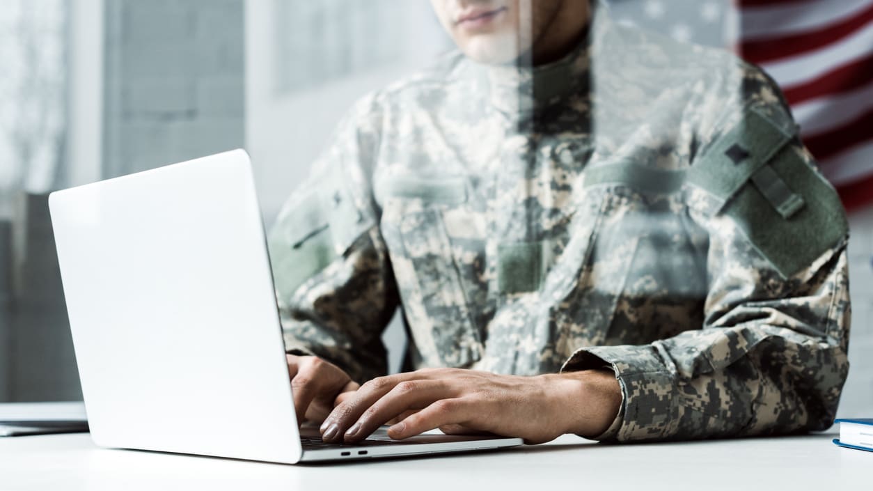 A soldier is using a laptop in front of an american flag.