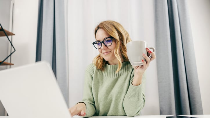 A woman wearing glasses is sitting at a desk with a laptop and a cup of coffee.