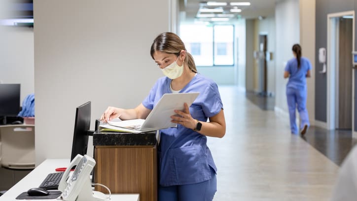 A nurse in scrubs standing at a desk in a hospital hallway.