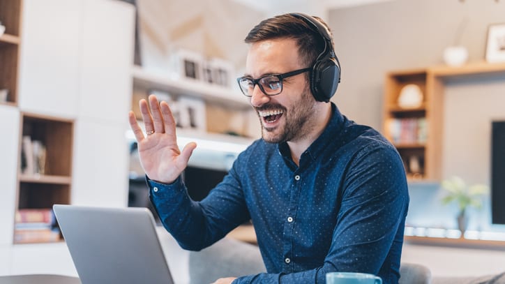 A man wearing headphones is using a laptop and waving at the camera.