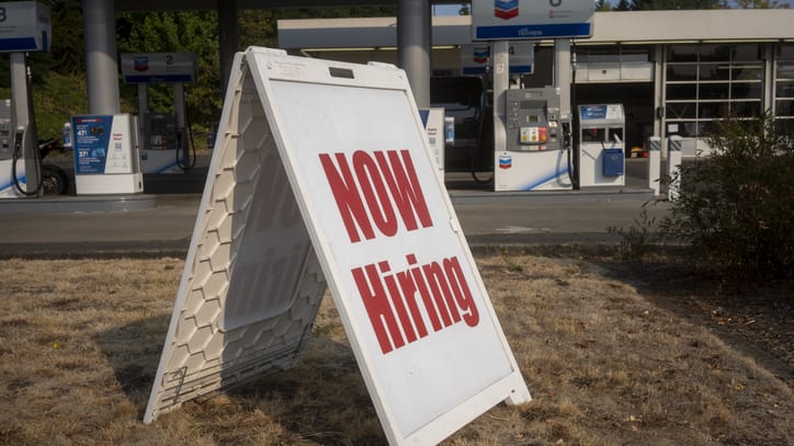 A sign that says now hiring in front of a gas station.