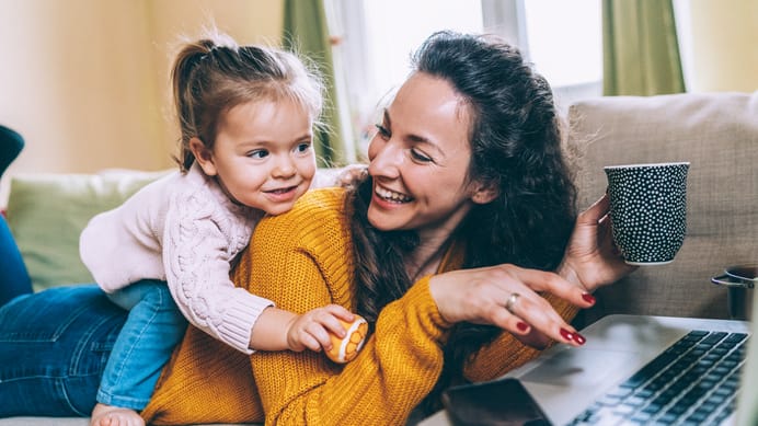 A mother and daughter sitting on a couch while using a laptop.