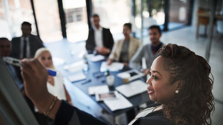 A business woman is writing on a whiteboard in front of a group of people.